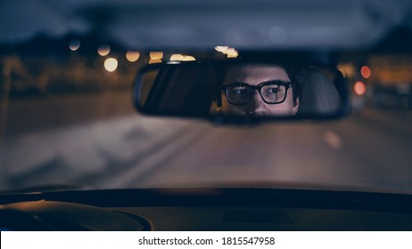 Driver's Face Is Reflected In A Rear View Mirror, Night Shot, Violet Color, Bokeh Light. Tired Concentrated Driver In Eyeglasses Is Reflected In A Mirror In Car, Night Road Shot Driving All Night