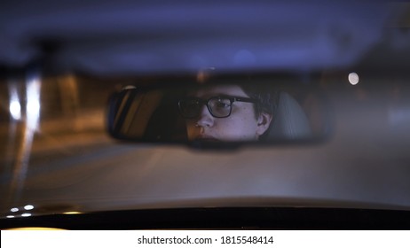 Driver's Face Is Reflected In A Rear View Mirror, Night Shot, Violet Color. Tired Concentrated Driver In Eyeglasses Is Reflected In A Mirror In Car, Night Road Shot Driving All Night