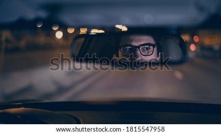Driver's face is reflected in a rear view mirror, night shot, violet color, bokeh light. Tired concentrated driver in eyeglasses is reflected in a mirror in car, night road shot driving all night