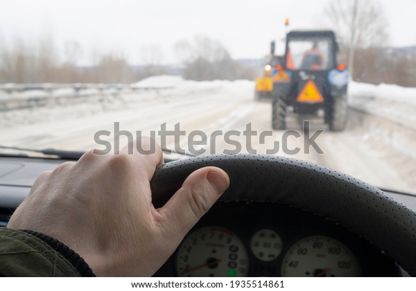 the driver\'s hand behind the wheel of a car\
that drives behind two slow-moving tractors that clear the road\
from snow in the cold winter\
season