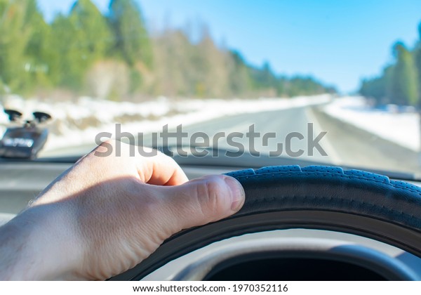 the driver\'s hand is on the steering wheel of a car\
that is driving on an asphalt road in sunny spring weather in the\
middle of a coniferous forest and on the side of the road lies\
white pure snow