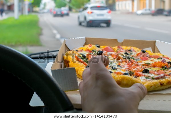 the driver\'s hand takes a piece of pizza,\
the packaging of which lies on the windshield panel of the parked\
car on the background of the city\
roadway