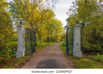 Driveway of an estate between an open black iron gate with stone pillars. The photo was taken on a cloudy day in the Dutch autumn season. The tree leaves are already changing color.
