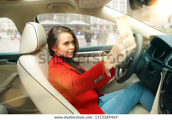 Driving around city. Young attractive woman driving\
a car. Young pretty caucasian model in elegant stylish red jacket\
making selfie photo at modern vehicle interior. Businesswoman\
concept. Human