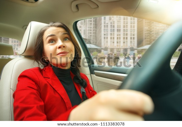 Driving around city. Young attractive woman
driving a car. Young pretty caucasian model in elegant stylish red
jacket sitting at modern vehicle interior. Businesswoman concept.
Human emotions concepts
