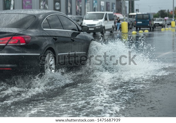  Driving car on\
flooded road during flood caused by torrential rains. Cars float on\
water, flooding streets. Splash on the car. Flooded city road with\
a large puddle