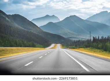 Driving down an empty road with a view of mountains in the distance and cloudy sky through a front car windshield.  - Powered by Shutterstock
