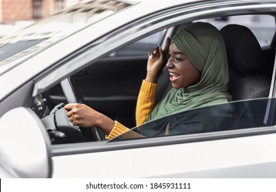 Driving Fun. Cheerful Black Muslim Woman In Hijab Listening Music And Singing In Car, Excited African Islamic Lady Enjoying Drive In City, Holding Steering Wheel And Looking On The Road, Side View