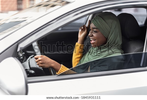 Driving Fun. Cheerful black muslim woman in hijab\
listening music and singing in car, excited african islamic lady\
enjoying drive in city, holding steering wheel and looking on the\
road, side view
