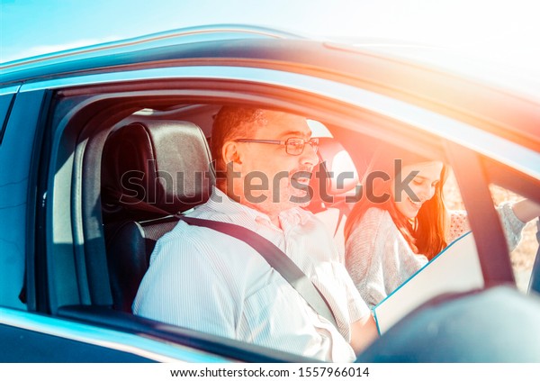 Driving male instructor taking notes on clipboard\
while sitting next to driving female teen student learning to drive\
on bright sunny day. Man tutor and teenage girl during driving\
class in the car