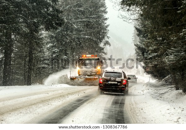 Driving on\
forest road through snowstorm, orange maintenance plough truck\
coming opposite way, view from car\
behind.