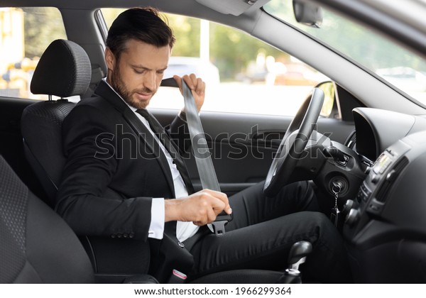 Driving safety\
concept. Handsome businessman fasten seat belt in his car, ready to\
go to office. Man in stylish suit putting on his seatbelt before\
driving car to airport, side\
view