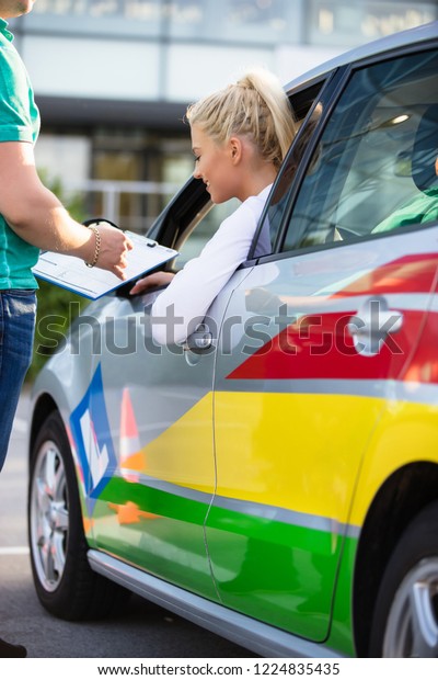 Driving\
school. Young woman or driving student on a driving test with her\
instructor. Learning to drive a car. Instructor of driving school\
giving exam while standing next to the\
car