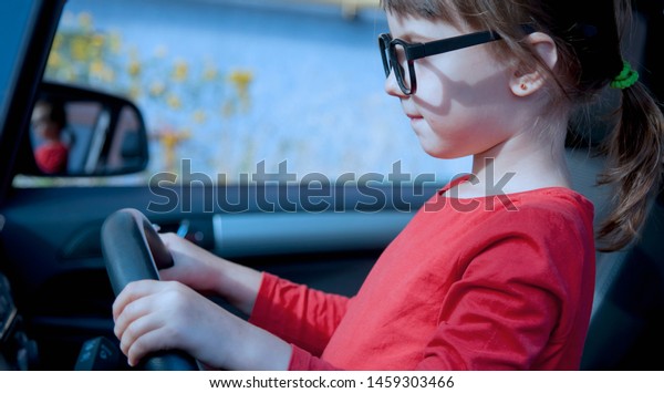 Driving shool. Cute little child\
girl holds wheel in a car and learns to drive. Humorous\
photo.