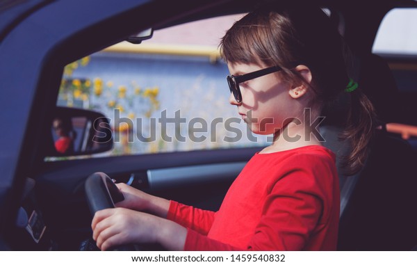 Driving shool. Humorous photo of\
cute little child girl holds a wheel in a car and learns to\
drive.