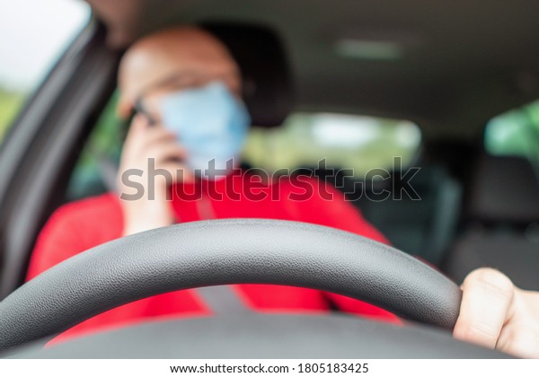 Driving wheel in focus, Bald\
driver out of focus using his smart phone while driving. Model in\
red shirt and blue medical face mask. Dangerous driving\
concept.