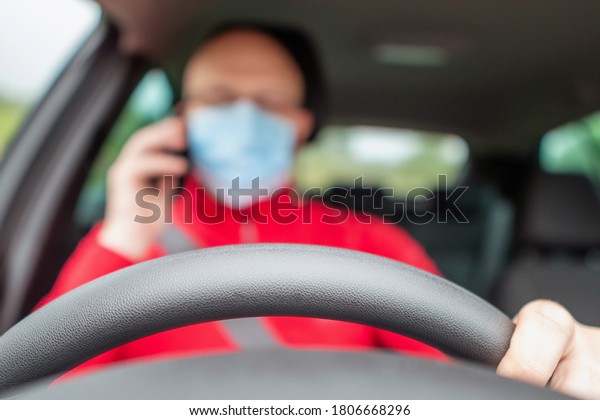 Driving wheel in focus, Bald\
driver out of focus using his smart phone while driving. Model in\
red shirt and blue medical face mask. Dangerous driving\
concept.