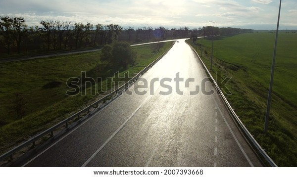Drone above wet asphalt road for cars between sown\
fields in spring morning. Wet asphalt road with reflective sun, sun\
glare. Wet road after rain between green fields. Aerial drone view\
flight