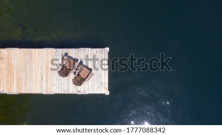 Drone aerial view of Two Adirondack chairs on a wooden dock overlooking the calm water of a lake in Muskoka, Ontario Canada.