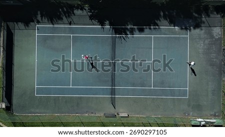 Drone captures two players practicing tennis on a hardcourt, showcasing focused training and athleticism. Crisp lines and shadows highlight their intense movements.