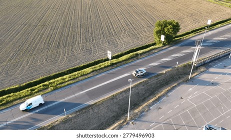 Drone capturing an expansive transportation network with asphalt road carrying two vehicles, an agricultural field showing furrows, and a large empty parking lot under sunlight