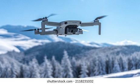 Drone with digital camera flying over winter landscape with snow covered mountains and trees. White remote controlled quadcopter hovering above snowy mountain peak. Close up shot of drone (UAV).