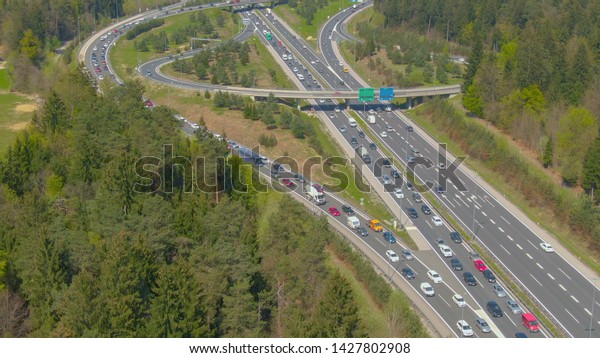 DRONE: Flying above a busy highway road network\
with overpasses and exits during peak rush hour in the hot sunny\
summer. Masses of tourists and commuters in cars occupy the freeway\
by the forest.