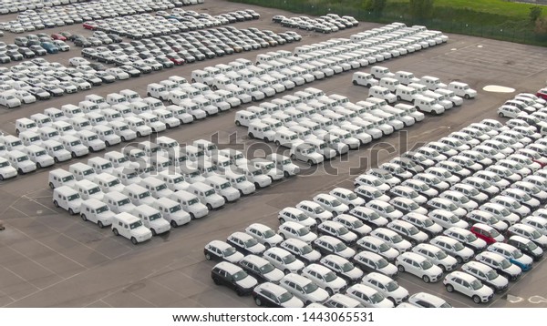 DRONE: Flying above a storage car park\
reserved for brand new imported cars. Countless cars wrapped in\
white paper are neatly organized in a massive parking lot serving\
as storage for car\
dealership.