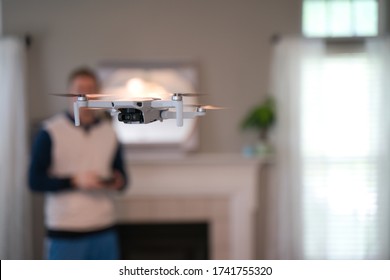 Drone Flying Indoors With Pilot Visible In Background. Amateur Drone Flight. User Wearing Sweatshirt Flying Drone Inside Of Home On Cold Day. White Drone Flying Inside.