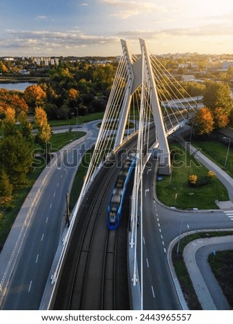 Drone forward overhead view of suspension bridge with tram movement in Krakow, Poland. Sunset top view of bridge structure and public transport in motion blur, empty asphalt road and park below