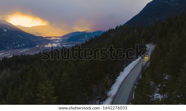 DRONE: Gorgeous orange sunset illuminates the\
forest as tourist car drives down a scenic highway running across\
British Columbia. Tourists on fun road trip drive down Sea to Sky\
Highway at sunrise
