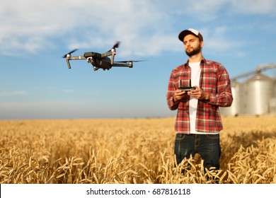 Drone Hovers In Front Of Farmer With Remote Controller Near Grain Elevator. Quad Copter Flies Near Pilot. Agronomist Taking Aerial Photos And Videos In A Wheat Field. Innovations In Agriculture.