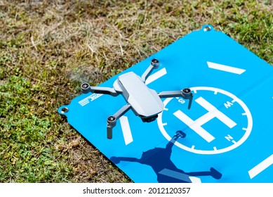 The drone hovers over the blue landing pad on the grass, the plastic propellers are on, and the camera on the gimbal is visible.