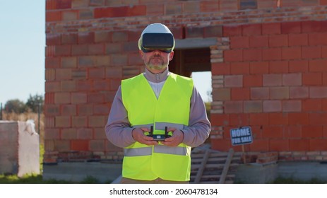 Drone Operator Controls An Aircraft, Holding Remote Controller. Professional Builder In Virtual Reality Helmet Standing In Front Of Construction Site. House Building. Business, Real Estate, Investment