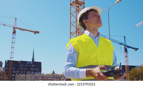 Drone operator holding remote controller. Professional builder in helmet and vest standing in front of construction site. Office building and crane background. Business, real estate and investment.