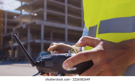 Drone operator holding remote controller. Professional builder in helmet and vest standing in front of construction site. Office building and crane background. Business, real estate and investment.