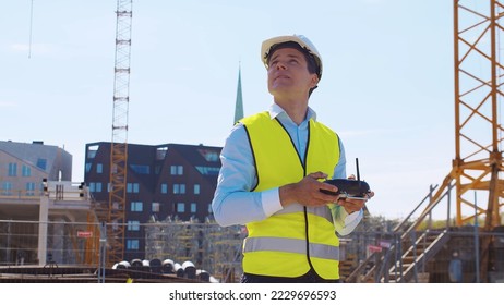 Drone operator holding remote controller. Professional builder in helmet and vest standing in front of construction site. Office building and crane background. Business, real estate and investment.