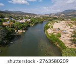 Drone perspective capturing the scenic Tecpan de Galeana river in Guerrero, Mexico. People enjoy swimming in its clear waters under a clear blue sky, surrounded by lush vegetation and distant mountain