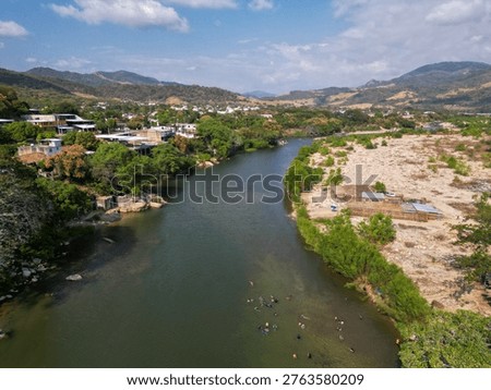 Drone perspective capturing the scenic Tecpan de Galeana river in Guerrero, Mexico. People enjoy swimming in its clear waters under a clear blue sky, surrounded by lush vegetation and distant mountain