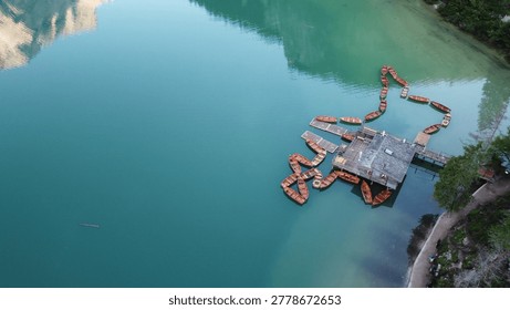 Drone perspective of modular floating platform and orange barriers on clear turquoise mountain lake, with shoreline path and forest edge.