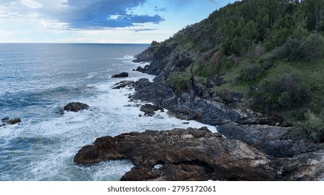 Drone perspective of ocean waves hitting a rugged rocky coastline with lush green vegetation.