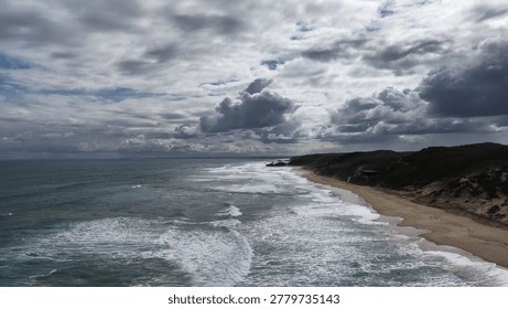 Drone perspective of white waves rolling onto a sandy shoreline beneath a moody overcast sky.
