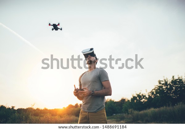 Drone\
pilot. Man using a drone with remote controller and virtual reality\
headset making photos and videos on sunset\
field