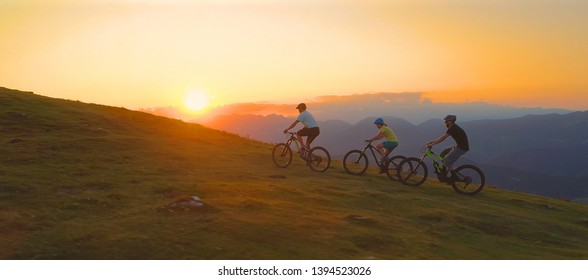 DRONE, SUN FLARE: Golden morning sunbeams shine on the active tourists pedalling bikes up a grassy hill. Three friends on a mountain biking adventure riding their bicycles up a mountain at sunset.