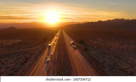 DRONE, SUN FLARE: Scenic shot of 18 wheeler trucks and cars crossing Mojave desert at dusk. Golden evening sun rays shine on the traffic moving up and down the straight freeway in rural California.