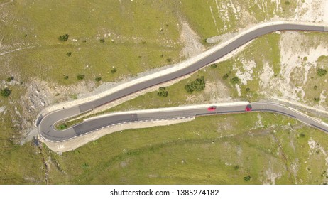 DRONE: Two red colored cars peacefully driving on a mountain pass in the stunning Italian Alps. Beautiful spring sun illuminates the picturesque landscape of the Dolomites on a perfect spring day