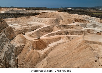 Drone view of an active open pit limestone quarry with terraced excavation levels, haul roads and heavy mining trucks. Industrial mining landscape showing raw material extraction and earthmoving operations.