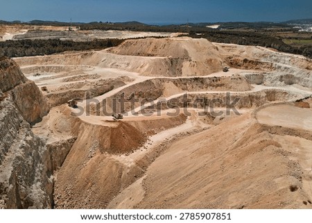 Drone view of an active open pit limestone quarry with terraced excavation levels, haul roads and heavy mining trucks. Industrial mining landscape showing raw material extraction and earthmoving operations.