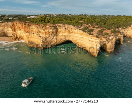 Drone view of Benagil Cave cliffs in Algarve, Portugal, with a tourist tour boat, limestone sea caves, turquoise Atlantic water, and waves.