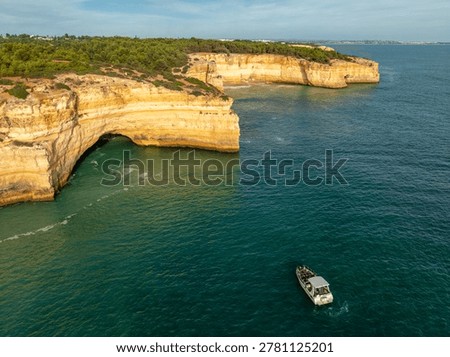 Drone view of Benagil Cave cliffs in Algarve, Portugal, with a tourist tour boat on Atlantic water, limestone sea caves, and rugged coast.